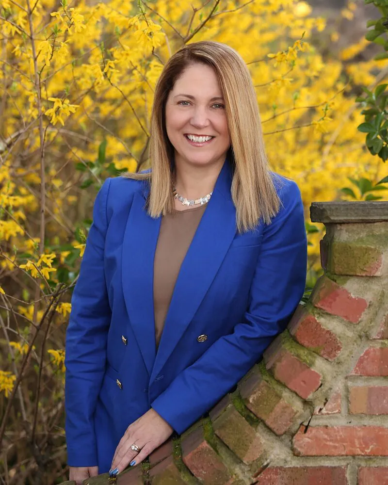 Jennifer Wilner with blonde hair leaning against red brick wall in bright blue blazer
